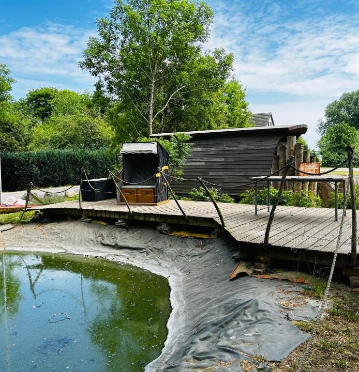 Ländliches Anwesen mit einem Teich, der von einem Holzsteg umgeben ist. Auf dem Steg stehen ein Strandkorb und ein kleiner Tisch.
