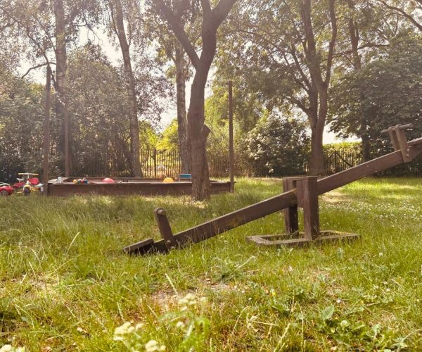 Spielbereich in einem großen Garten mit einer hölzernen Wippe auf einer Wiese. Im Hintergrund sind ein großer Sandkasten mit Spielzeug und mehrere Bäume zu sehen.