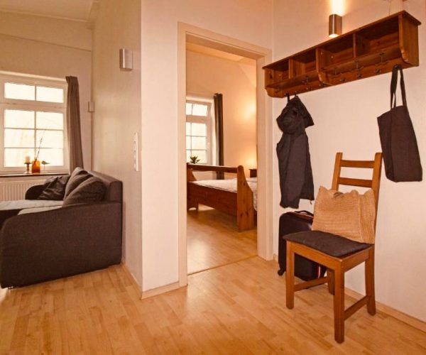 Eingangsbereich einer Ferienwohnung mit einem rustikalen Holzgarderobensystem, an dem Jacken hängen, und einem Stuhl mit Koffer.