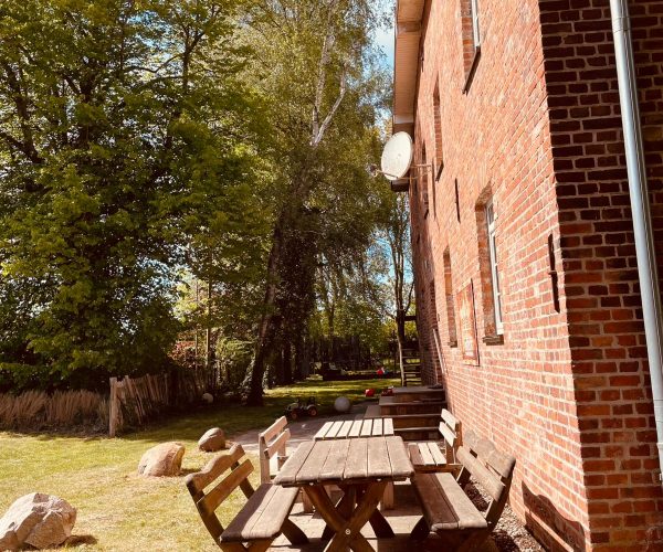 Eingangsbereich eines historischen Backsteingebäudes mit einem rustikalen Holztisch und zwei Bänken auf einer gepflasterten Terrasse direkt an der Fassade.