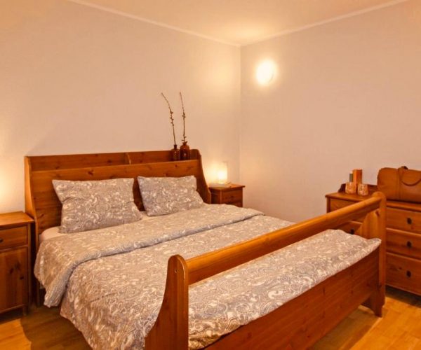 Schlafzimmer in einer Ferienwohnung mit einem großen, rustikalen Holzbett und hellen Nachttischen und einer Kommode. Das Bett ist mit einer grau-weißen, gemusterten Bettwäsche bezogen.