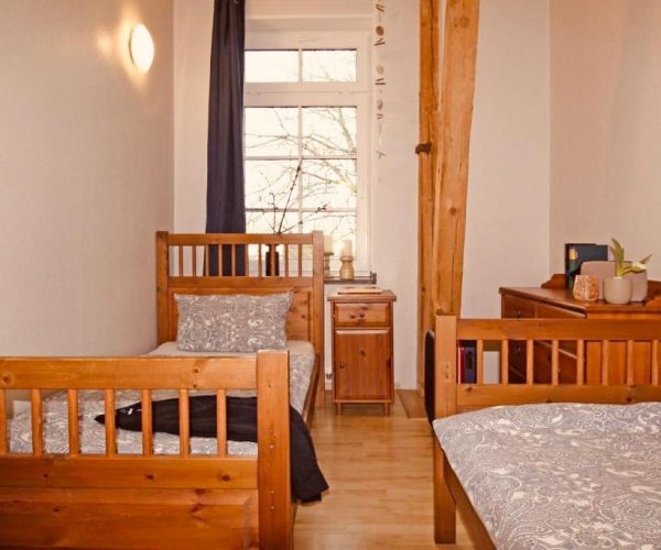 Schlafzimmer in einer Ferienwohnung mit zwei rustikalen Einzelbetten aus hellem Holz, die mit grauer Bettwäsche bezogen sind.